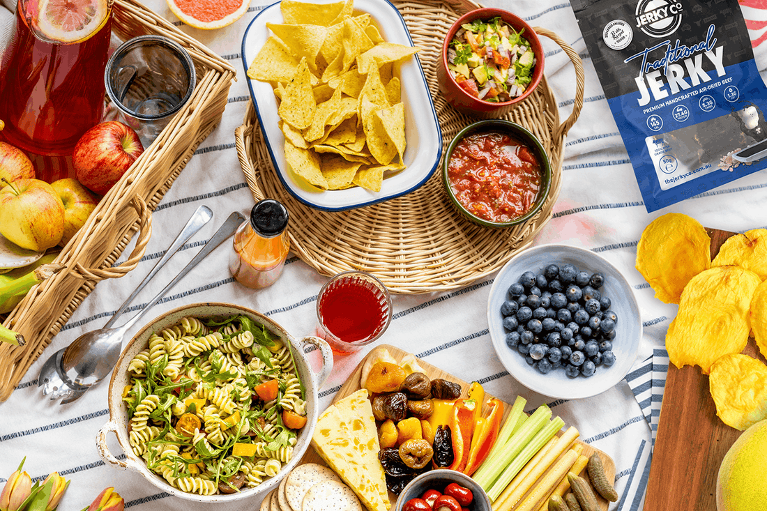 Summer picnic spread with fresh fruit, crackers, cheese, vegetables and dips, with space for The Jerky Co air-dried fruit and meat snacks.